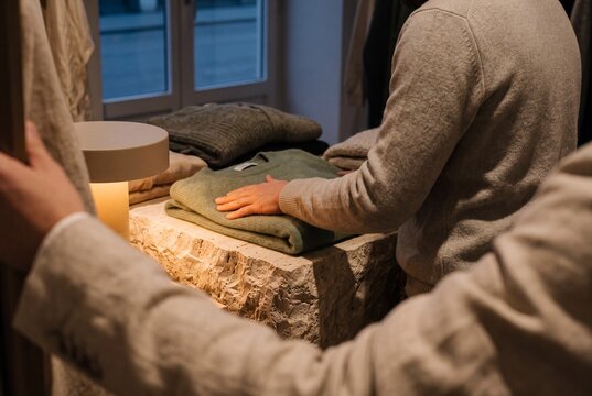 A customer touches a folded soft wool sweater displayed on a stone table in a modern luxury fashion boutique