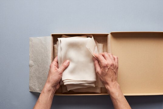 Hands Unpacking Folded Silk Fabric from a Cardboard Gift Box with Tissue Paper on a Neutral Background