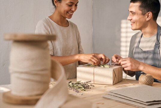 Smiling young couple wrapping a gift box using eco-friendly craft paper and dried lavender in a sunny workshop