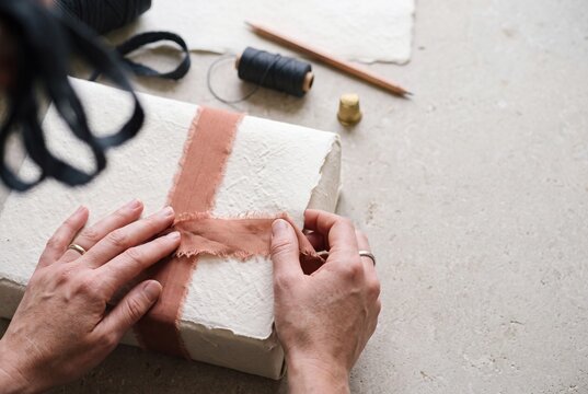 Hands wrapping a gift box using handmade textured paper and frayed terracotta cloth ribbon on a neutral craft table