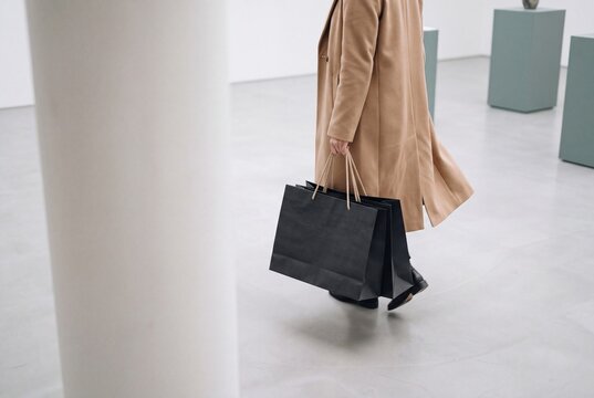 Stylish woman in a beige coat carries black paper shopping bags while walking through a minimalist interior