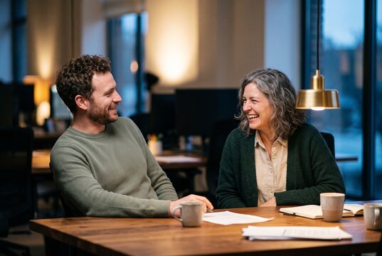 Two cheerful diverse colleagues discussing business ideas while laughing together at wooden office desk during a meeting