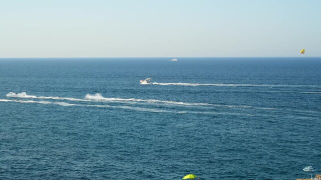 Water scooters racing across deep blue sea leave long white spray trails near the horizon, while a yellow parasail floats in the sky and beach umbrellas sit on shore, concept of summer water sports a