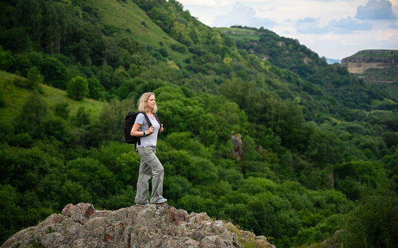 Caucasian woman with backpack standing on a rock. Female looking at green mountain landscape near Kislovodsk, Russia. Outdoor adventure, hiking and active lifestyle concept