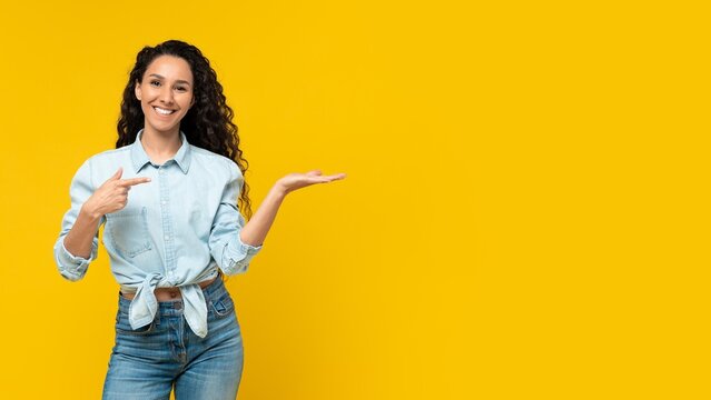 Smiling Young Hispanic Woman Pointing With Hand In Front Of Yellow Background, Showcasing Product Or Service, Energetic And Happy Emotion, Ideal For Advertising Or Marketing Purposes.