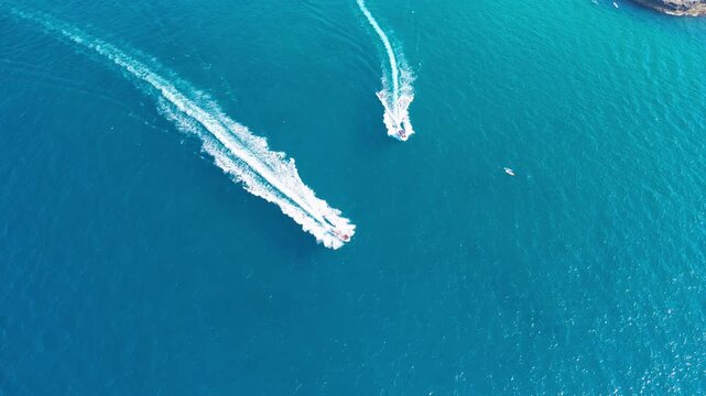 Speedboat carving a curved white wake across turquoise sea as a second boat trail cuts diagonally below. Rocky shoreline peeks in from the upper corner, concept of speed, motion and summer water spor