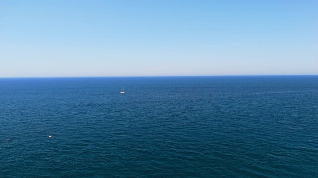 Under cloudless blue sky, calm deep blue sea stretches to a straight horizon with a tiny sailboat in the distance and small buoys floating in the foreground, concept of peace, freedom and minimalist 