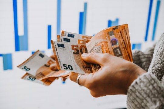 Hands holding euro currency in front of the stock exchange screen showing trading graphics.