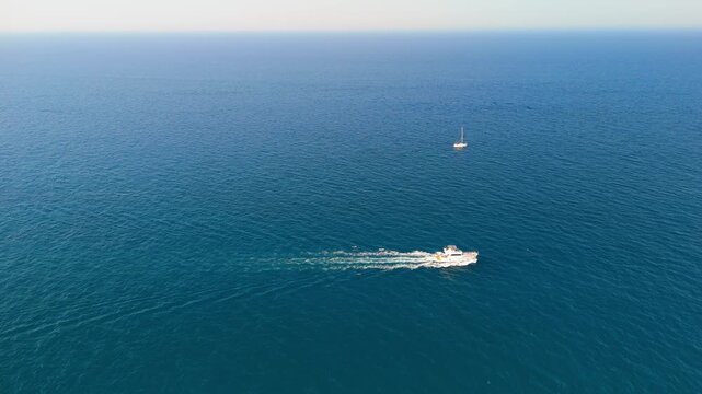 White tour boat sailing alone across vast deep blue sea, leaving a long foamy wake behind it while a small vessel drifts near the hazy horizon, concept of sea journey, solitude and summer excursions