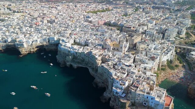 Small boats floating at anchor in a dark bay below the dense whitewashed rooftops of Polignano a Mare, the town spreading far inland from its cliff edge, concept of mediterranean urban density and se