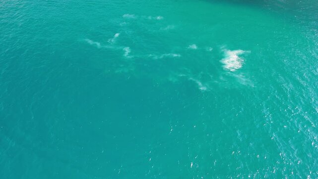Top down view of turquoise sea with scattered white foam patches drifting on the surface after a passing boat, sunlight sparkling across the rippled water, concept of calm summer seascape and natural