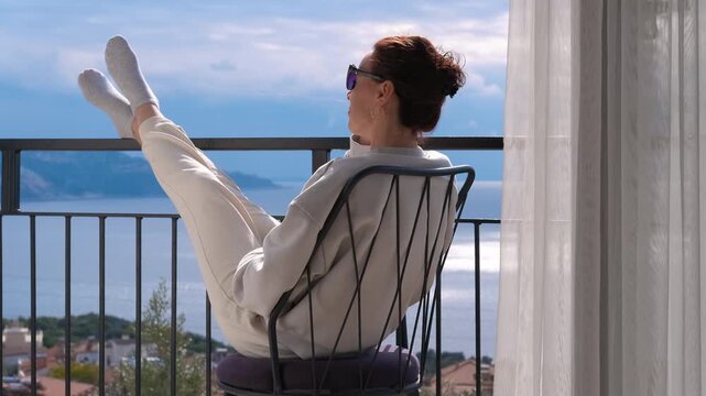 Woman relaxing on a balcony enjoying the sea view. Calm woman with her feet up relaxing in a chair on her balcony, peacefully enjoying the beautiful sea view and the fresh air during tranquil morning