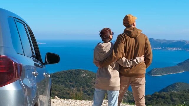 Romantic couple hugging and enjoying sea view during road trip. Loving couple standing on a mountain viewpoint, hugging and admiring the beautiful blue sea and islands below after arriving by car