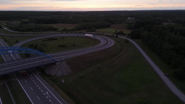 Trucks and cars driving around a looping highway exit ramp surrounded by dark fields, streetlights lining the curve under a faint pink glow on the horizon, concept of freight logistics, road travel a