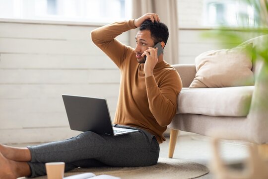 Worried Black Man In His 30s Sitting On Floor, Taking Phone Call While Using Laptop At Home Office, Appearing Stressed And Concerned About Work. Focused On Troubleshooting Business Issues In