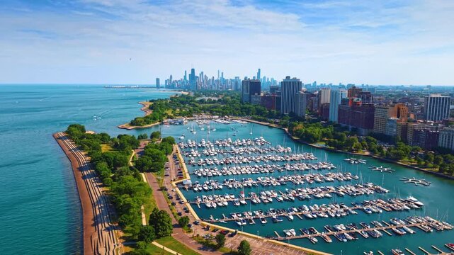 Belmont Harbor boat rows and Chicago lakefront from above. White yachts fill parallel docks beside a stepped seawall, trees and apartment towers in sunlight.