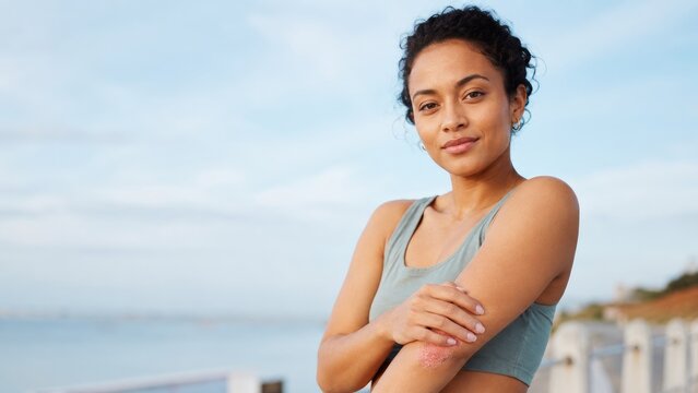 Young woman in a sports top by the sea, smiling confidently with arms crossed while showing skincare on her elbow
