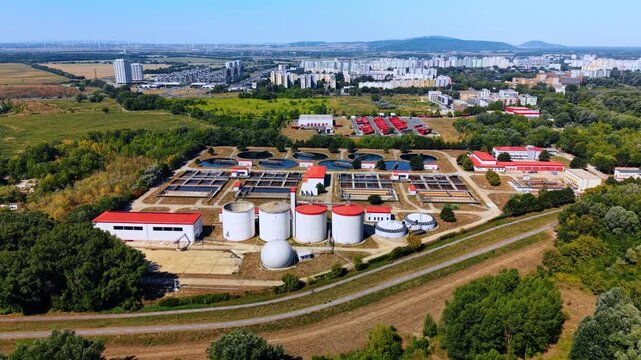 Large white sludge digesters at water treatment plant in Bratislava. Panoramic view displays round storage silos and red structures near green forest.