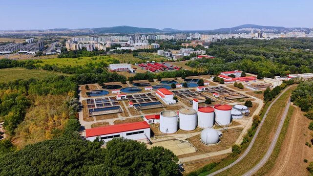 Wastewater treatment plant beside woodland and parallel paths. An oblique aerial view shows tanks and basins, with a bus depot and residential blocks in the background.