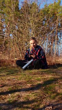 Young adult woman sitting on ground in woodland holding smartphone for video call or selfie while notebook and stainless steel thermos rest nearby. Smiling expression in sunny autumn landscape with