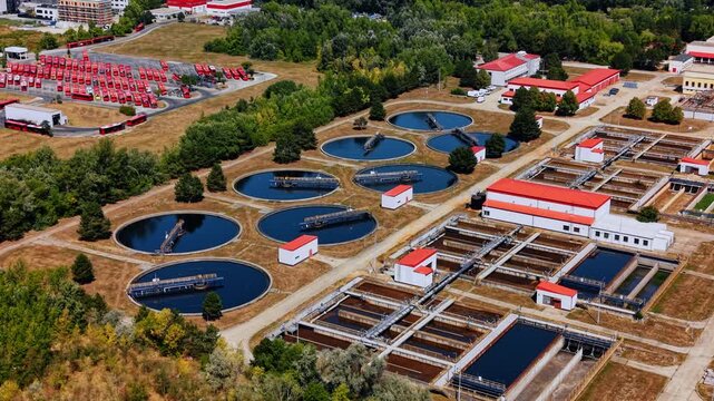 Rows of wastewater clarifiers beside rectangular treatment basins. Metal bridges span blue water tanks, while pipes cross brown basins near buildings and a bus depot.