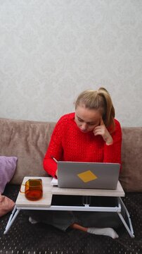Young woman sits comfortably on sofa using laptop on portable lap desk at home. Relaxed home workspace includes notebook mug, tablet and soft furnishings in cozy interior. Suitable for remote work