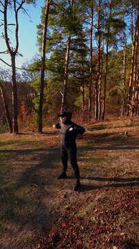 Young adult woman performs side lunge stretch during outdoor fitness session. Person wears black hoodie, cap, sweatpants and boots while warming up on grassy ground surrounded by autumn trees in