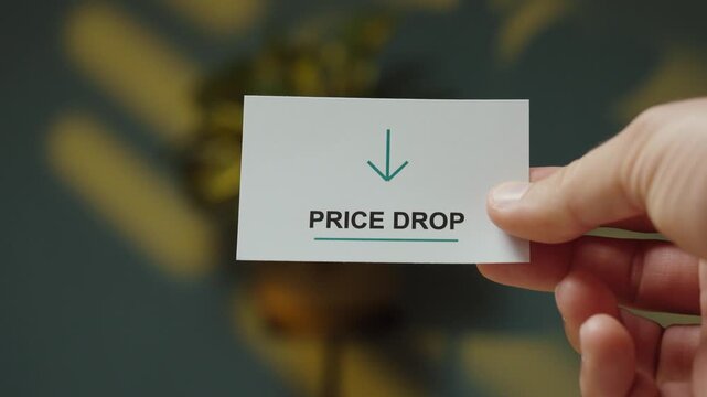Close-up of a hand holding a &ldquo;Price Drop&rdquo; card with downward arrow, representing falling prices, discounts, lower costs and market changes.