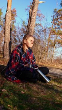 Young adult woman sitting on grass in forest clearing while writing in notebook, wrapped in plaid blanket beside stainless steel thermos. Calm focused moment in sunny autumn nature with warm light
