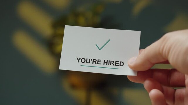 Man flips a small card to reveal a &ldquo;You&rsquo;re Hired&rdquo; message with a check mark, representing successful recruitment and getting a new job.