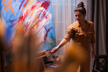 Artist pauses beside a colorful abstract canvas and looks down at a wide brush in hand, a quiet moment within the creative painting process, highlighting observation and artistic expression. © DC Studio