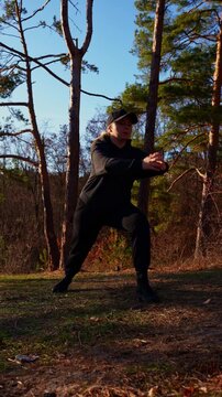 Young adult woman performs side lunge stretch during outdoor fitness session. Person wears black hoodie, cap, sweatpants and boots while warming up on grassy ground surrounded by autumn trees in