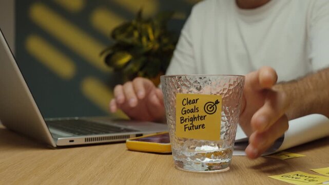 Man takes a sip of water, puts the glass with a &ldquo;Clear Goals Brighter Future&rdquo; sticky note back on the desk and continues working on his laptop.