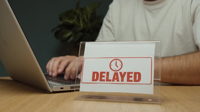 Man works on his laptop with a &ldquo;Delayed&rdquo; sign on the desk and checks his watch, representing waiting, schedule delays and time pressure.