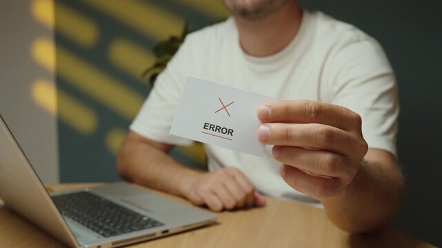 Man flips a small card and holds the &ldquo;Error&rdquo; message with a red cross toward the camera, representing a system error, technical problem and failed digital process.