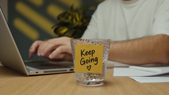 Man takes a sip of water from a glass with a &ldquo;Keep Going&rdquo; motivational sticky note while working at his laptop.