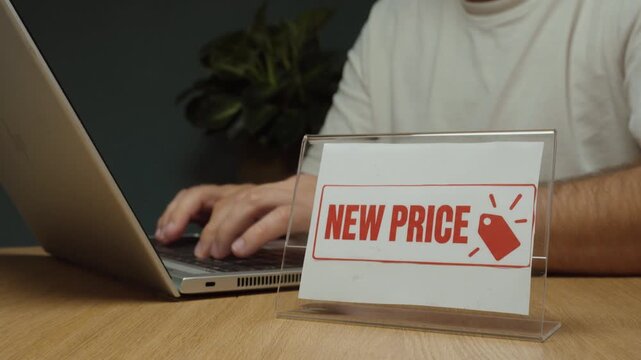 Man works on a laptop and then places a &ldquo;New Price&rdquo; sign on the desk, representing price changes, retail pricing, sales and updated costs.