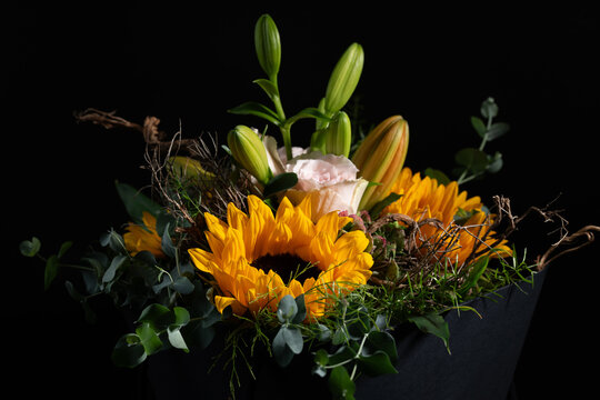 A close-up of a bouquet of flowers in the fall. Sunflowers, roses, and lilies glow in warm fall colors against a dark background.