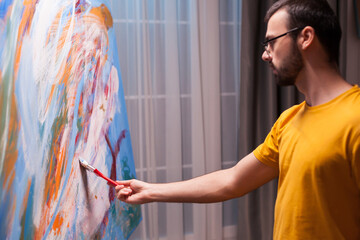 Male artist uses a brush to build color and texture across the lower area of a large abstract canvas during focused studio work and an evolving creative process, highlighting concentration. © DC Studio