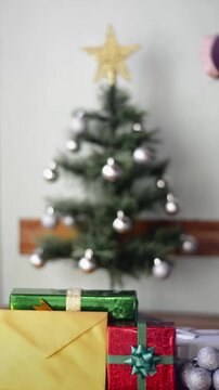 Wrapped gifts, envelope and silver balls in sharp focus in foreground with male hands placing Christmas lights on tree blurred in the background