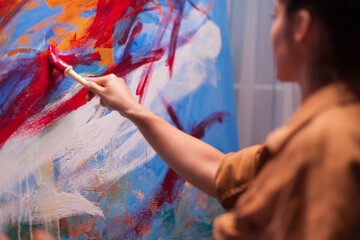 Close framing shows a brush spreading thick red paint across an abstract canvas as the woman artist works just out of focus, revealing texture, motion and hands-on technique, using selective focus. © DC Studio