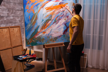 Male artist applies a bold orange brushstroke across a large abstract canvas, combining energetic color, hands-on creativity and expressive studio painting, highlighting artistic expression. © DC Studio