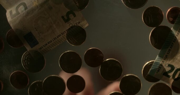 Low-angle view from beneath a glass table showing an unrecognizable person moving and counting euro coins by hand. The conceptual scene represents personal finances, taxation, poverty, social benefits