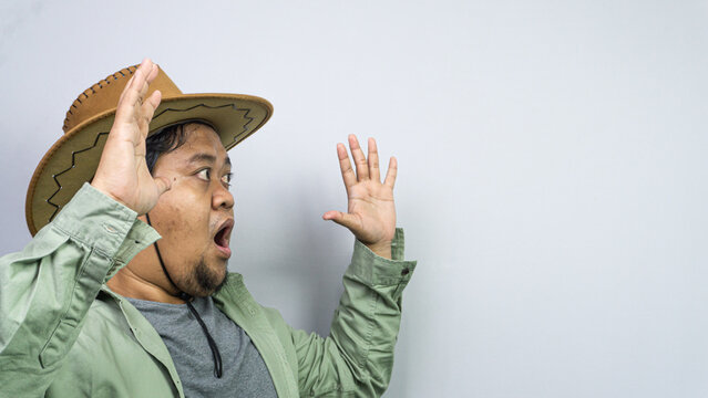 A completely shocked adult Asian male wearing one brown western hat and casual green shirt raises both hands against a plain grey background. That terrified individual demonstrates