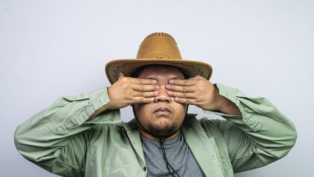 Experiencing overwhelming fear or willful ignorance, a young Asian male covers both eyes tightly using flat hands against some plain light background. The subject sports western headwear