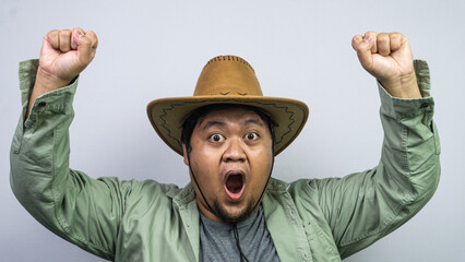 A young Asian male wearing a brown cowboy hat plus some casual green clothing stands against plain light background with arms raised in triumphant gesture. The person features an open © M. Faisal Riza
