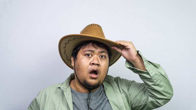 Executing a classic greeting gesture, one young Asian male tips the front brim of his brown cowboy hat while standing against plain light background. The individual displays slightly