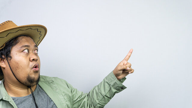 A young male wearing a brown cowboy hat looks upwards and points his index finger to the right with an expression of sudden realization or mild surprise against a light gray background.