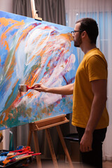 Artist wearing glasses adds careful brushwork to a large colorful abstract canvas, combining concentration, creative craft and precise hands-on painting in the studio, highlighting detailed brushwork. © DC Studio