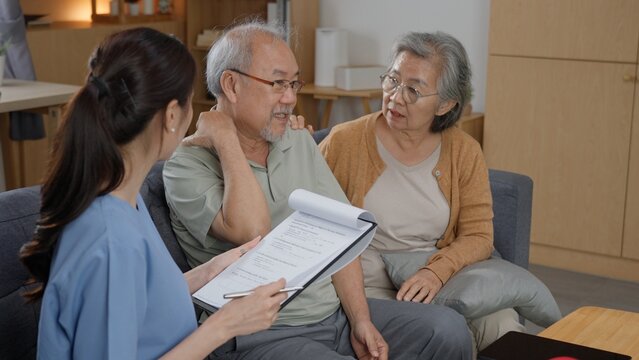 Asian female nurse holding medical clipboard and pen while consulting senior man with neck pain sitting with his caring wife during healthcare home visit in cozy living room.